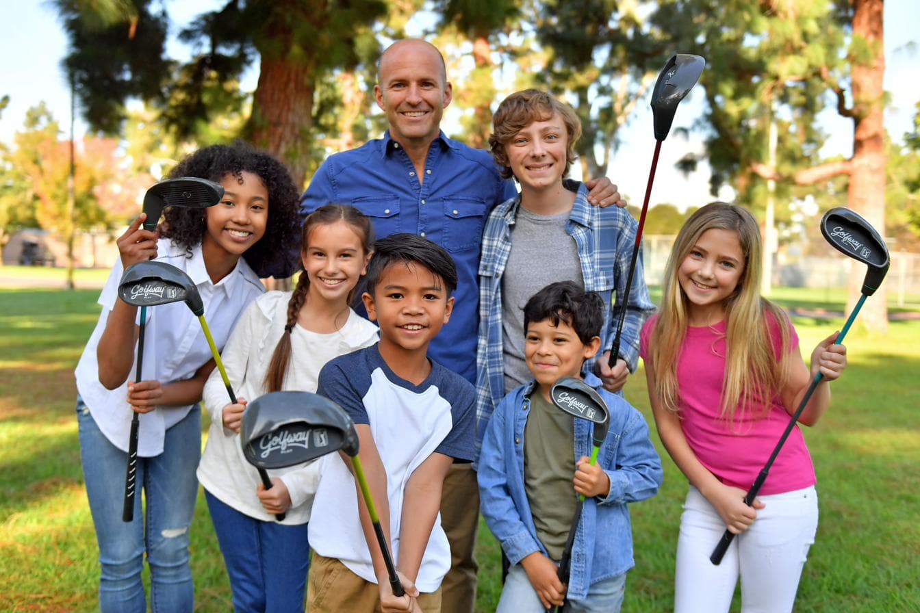An adult and six smiling children holding junior golf clubs on a school field.