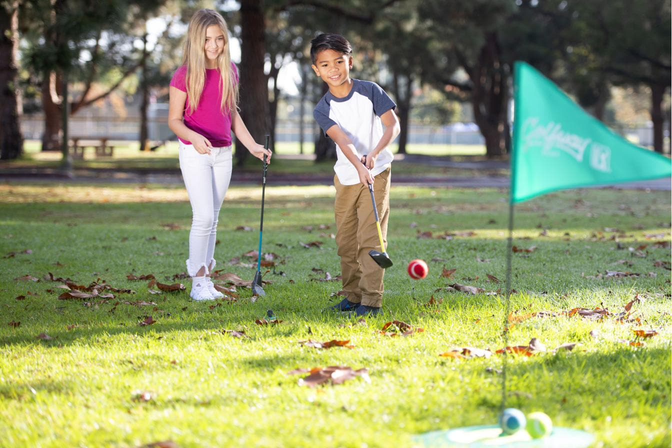 Two children chipping a ball towards a flag target on a grass field.